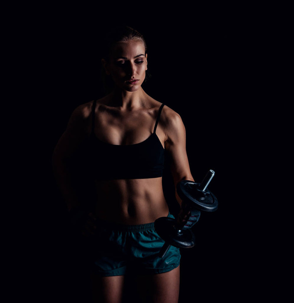Portrait of a young fitness woman in sportswear doing workout with dumbbells on black background. Tanned sexy athletic girl. A great sporty female body. - Fotó, kép