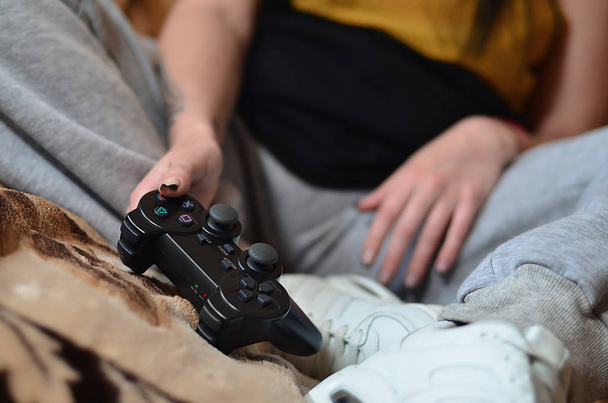 Un fragmento del cuerpo de una joven caucásica que se sienta en el sofá y juega videojuegos con un joystick negro moderno. Imagen de primer plano de un controlador de juego en manos femeninas
 - Foto, imagen