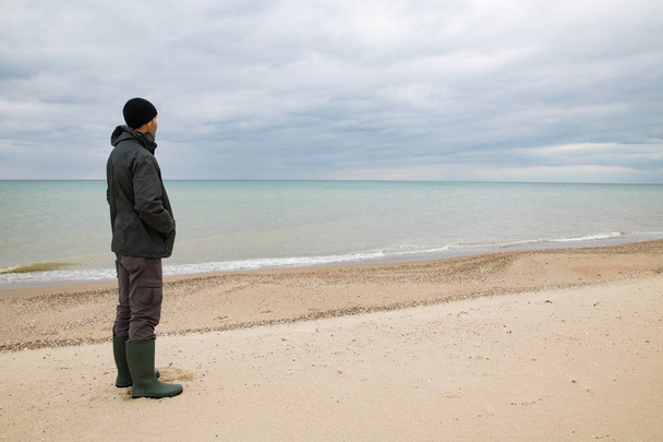 Mann in grüner Jacke und kniehohen Stiefeln steht am Strand und blickt aufs Meer. grünes Meerwasser, Gewitterwolken im Hintergrund - Foto, Bild