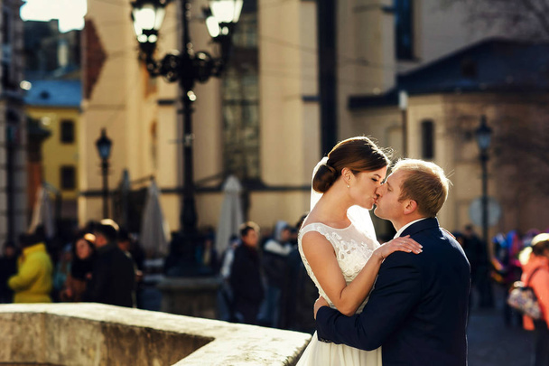 Groom kisses a bride standing in the sunrays on a citysquare - Φωτογραφία, εικόνα