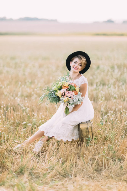 The vertical portrait of the smiling bride with the colourful bouquet sitting on the vintage suitcase in the spring field. - Фото, зображення