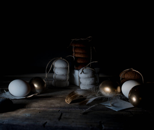 Easter. Easter night. Golden eggs and cakes on a wooden table. White feathers. Vintage. Dark background - Photo, image