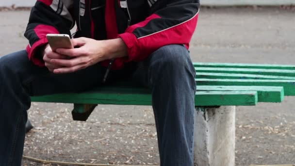 スマート フォンで何かを入力するベンチの公園で座っている孤独な男 - 映像、動画