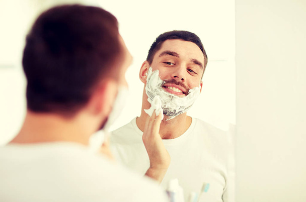 happy man applying shaving foam at bathroom mirror - Foto, imagen