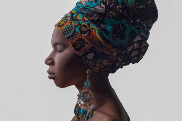 Young beautiful African woman in traditional style with scarf, earrings crying, isolated on gray background. racism, depression or loneliness concept - Фото, изображение