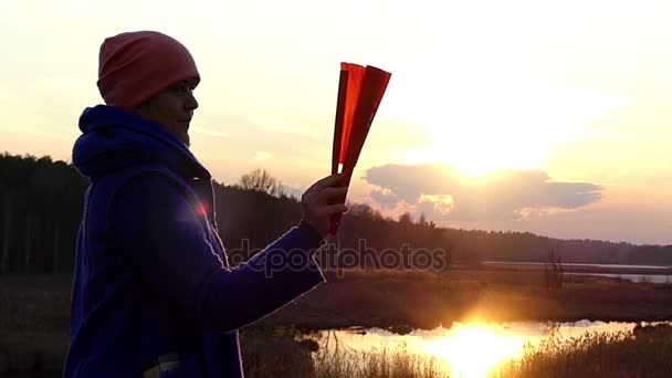 Prachtige vrouw ontvouwt zich een oranje Handheld Fan onder de stralen van een zonsondergang, de zon in het vroege voorjaar in Oost-Europa - Video