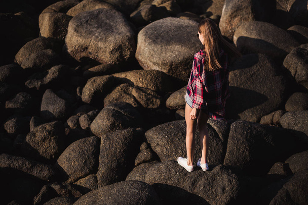 Krásná štíhlá žena v košili, šortky a módní sluneční brýle chodit na velké kameny. Pohled z zpět. Krása roztomilé děvče na břehu oceánu moře tropické pláže s velkými kameny. Letní venkovní životní styl. - Fotografie, Obrázek
