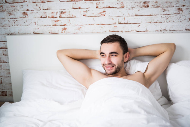 schöner gut gebauter Mann, der sich morgens in seinem Bett ausruht, mit einem Lächeln an der Ziegelwand im Zimmer zu Hause - Foto, Bild