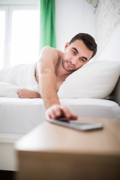 Мужчина в постели просыпается от тревоги на мобильном телефоне утром
 - Фото, изображение