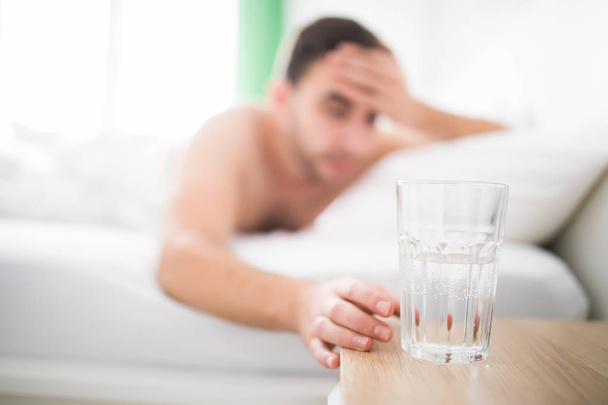 Homem bonito acordar com dor na cabeça sofre de dor de cabeça e estender-se para obter um copo de água em sua cama
 - Foto, Imagem