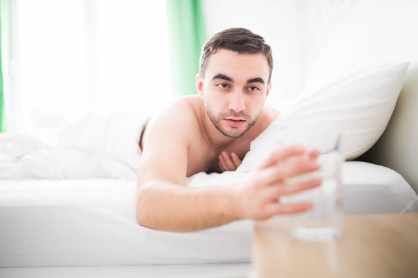 Красивый молодой человек просыпается утром и протягивает, чтобы выпить стакан воды в постели
 - Фото, изображение
