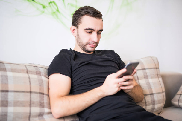 Cheerful handsome young man using his smart phone while sitting on the couch at home - Foto, Bild