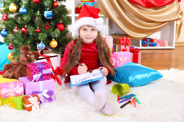 Beautiful little girl in red dress writes letter to Santa Claus in festively decorated room - Zdjęcie, obraz