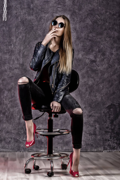 Beautiful urban trendy girl in black leather jacket and jeans posing on a high chair - Foto, Bild