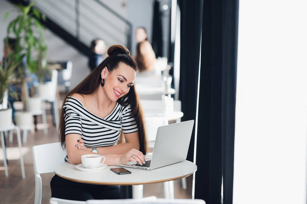 Image of happy woman using laptop while sitting at cafe. Young woman sitting in a cafe and talking with friends through the internet. - Photo, Image