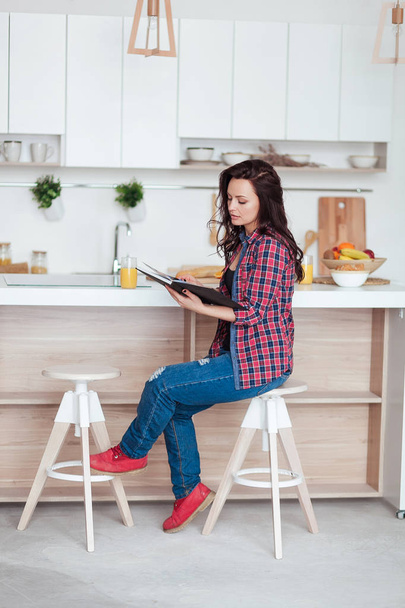 Завтрак - Улыбающаяся женщина читает книгу на белой кухне, свежий апельсиновый сок
 - Фото, изображение