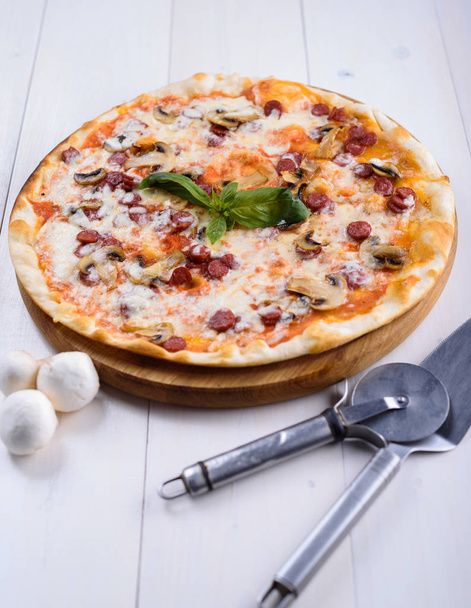 Pizza with mushrooms and hunting sausages on a wooden board next to the devices for pizza - Photo, image