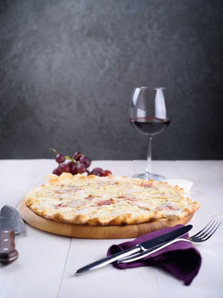 Pizza bianca with chicken, ham and cream sauce on a wooden board next to a glass of wine, grapes and pieces of cheese - Fotó, kép