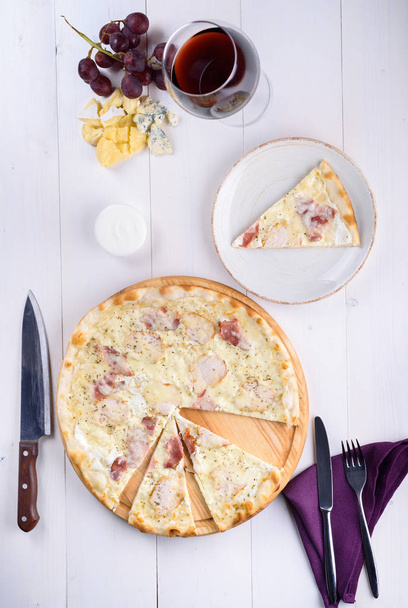 Pizza bianca with chicken, ham and cream sauce on a wooden board next to a glass of wine, grapes and pieces of chees - Fotó, kép