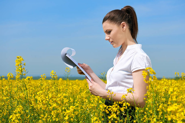 Деловая женщина в цветочном поле на улице смотрит на буфер обмена. Молодая девушка на желтом поле рапса. Прекрасный весенний пейзаж, яркий солнечный день
 - Фото, изображение