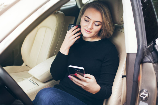 Kaunis nuori nainen istuu autossa kuljettajan istuimella ja juo kahvia ja katselee matkapuhelinta.
. - Valokuva, kuva