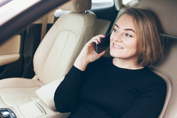 Close-up portret van een mooie gelukkig en lachen met alle tanden jonge Kaukasische vrouw die in de auto zitten en praten over haar smartphone. - Foto, afbeelding