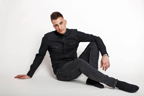 Stylish handsome man in black shirt and pants sits in the studio on a white background - Фото, изображение