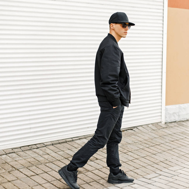 Hombre guapo en gafas negras y una gorra de béisbol negra, chaqueta de moda y zapatillas de deporte caminando por la calle
 - Foto, Imagen