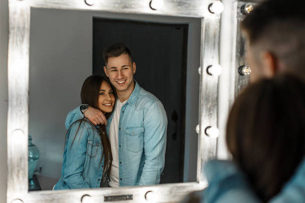 Beautiful happy young couple with a smile in stylish denim clothes hugging near a vintage mirror with lights - 写真・画像
