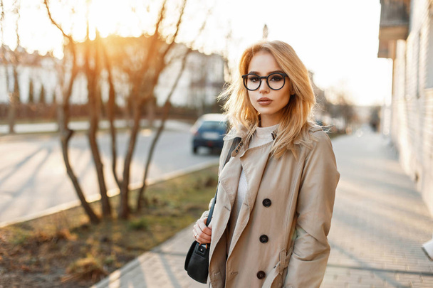 Beautiful young woman in stylish glasses and trendy coat with handbag in the day of spring at sunset - Photo, Image
