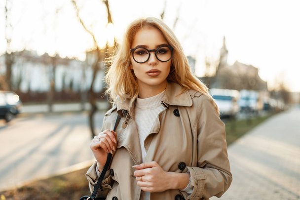 Beautiful stylish young woman with trendy glasses in coat with bag walks in the spring at sunset - Foto, Imagen