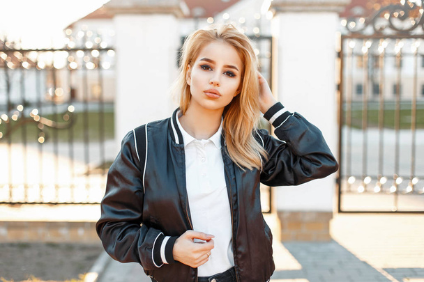 Modieuze jong blond meisje in een zwarte jas en witte polo shirt in een lentedag - Foto, afbeelding