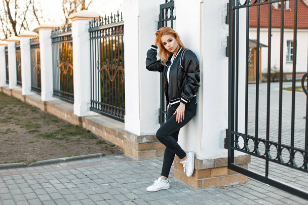 Bir bombardıman ceket siyah kot ve beyaz ayakkabı ile güzel genç kadın kolej çit duruyor - Fotoğraf, Görsel