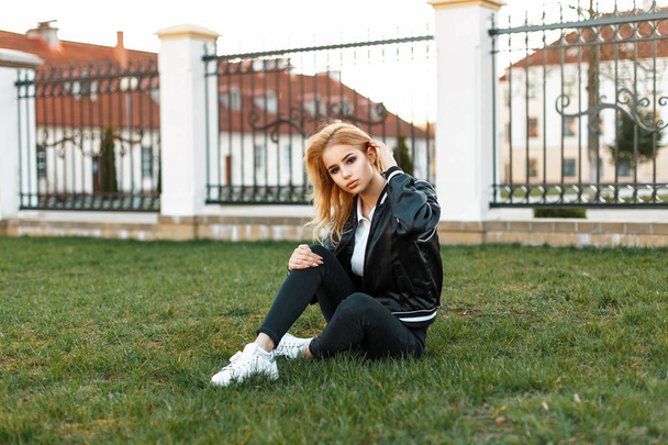 Jonge mooie vrouwelijke student in modieuze jas, zwarte jeans en witte sneakers zit op gras - Foto, afbeelding