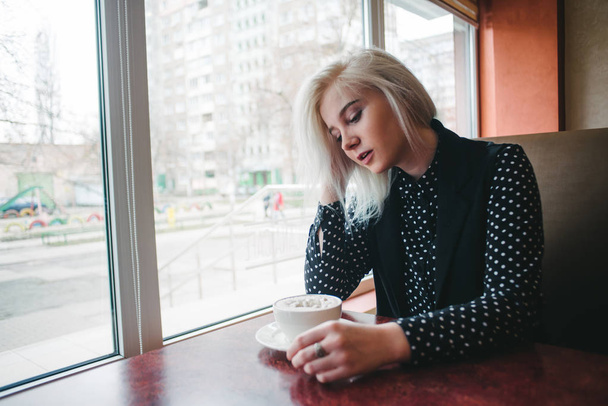 Porträt einer jungen schönen blonden Frau, die mit einer Tasse Kaffee in einem gemütlichen Café am Fenster sitzt. Kaffeepause - Foto, Bild