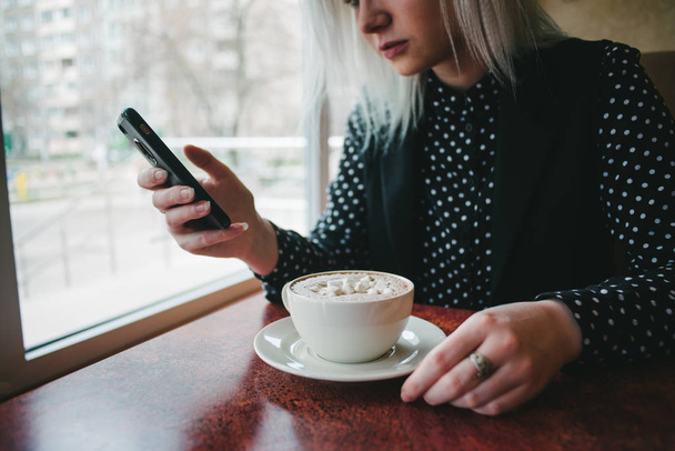 Молодая женщина в черном пользуется телефоном в кафе и пьет кофе с зефиром. Перерыв на кофе в кафе
 - Фото, изображение