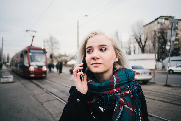 junge schöne blonde Mädchen trägt warm, dass am Telefon auf dem Hintergrund der städtischen Landschaft sprechen. Außenporträt auf dem Hintergrund der Straßenbahn - Foto, Bild