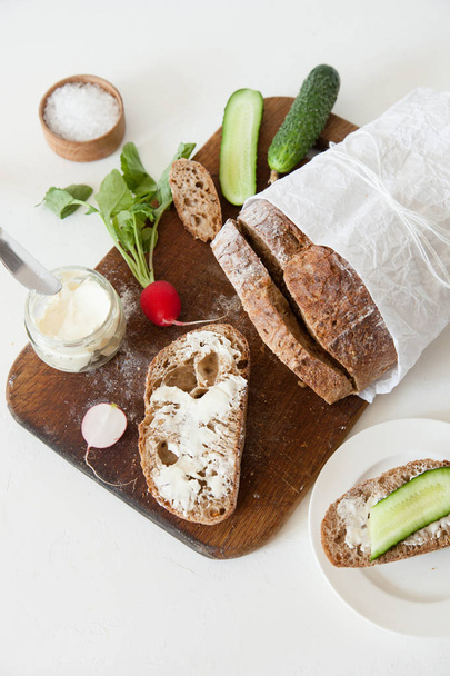 Brood met zout, boter, komkommer en radijs leugen op een houten snijplank op een witte achtergrond. - Foto, afbeelding