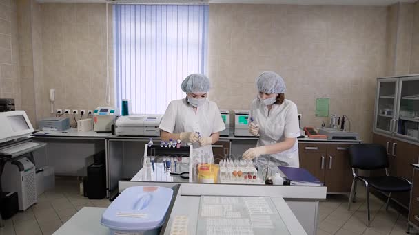 zwei Krankenschwestern in weißen Mänteln überprüfen Röhrchen mit einem Bluttest von Patienten, um die Forschung unter dem Mikroskop durchzuführen oder legen Glasprobenröhrchen in eine Krankenhauszentrifuge - Filmmaterial, Video