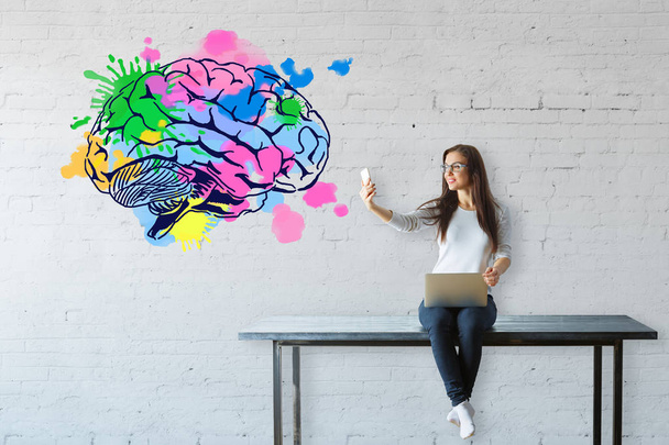 Young woman sitting on table, using laptop and taking selfie with smartphone on white brick background with colorful brain sketch. Brainstorm concept - Photo, Image