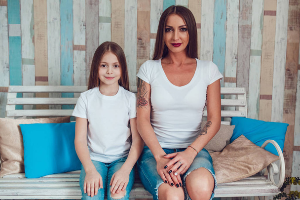 Mom and daughter with beautiful long hair in identical clothes are sitting on a wooden couch. Blank white T-shirt. - Foto, Imagem