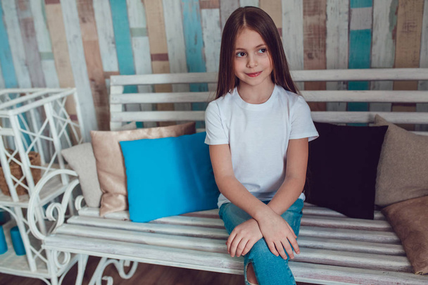 Beautiful little girl in blank white T-shirt and jeans is sitting on a wooden couch. - Фото, изображение