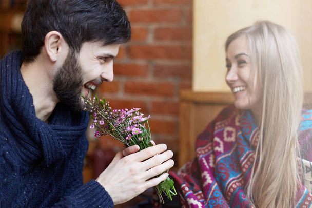 Focus on man who is taking flowers from a young attractive lady. A pair of lovers is laughing in a cozy cafe, having good time and enjoying each other.  - Foto, Imagem