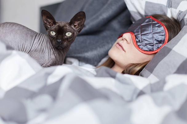 Close up retrato de menina e esfinge gato. Ambos em foco. Jovem bela senhora está dormindo usando máscara de dormir em seu quarto. Pet está observando de lado carregando seus sonhos em seu peito
. - Foto, Imagem
