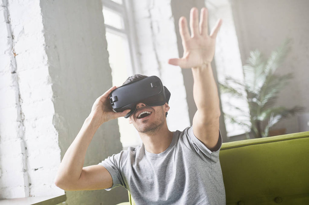 Hansome european male experiencing virtual reality using VR headset glasses, sitting on couch at home and throwing hand in air, gesturing. Young  man wearing oculus or goggles playing video games - Photo, image