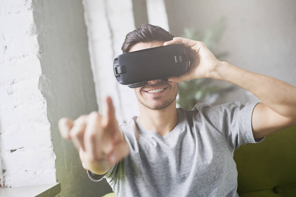 Closeup portrait of european attractive man pointing with finger as if interacting with something, wearing virtual reality goggles in modern coworking studio. Future is now. Using VR technology - Foto, Bild