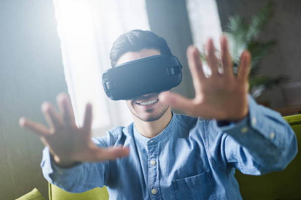 Closeup portrait of astonished handsome guy wearing blue denim shirt experiencing virtual reality while using oculus rift headset for entertaining, sitting on couch in coworking studio. Lens flares  - Foto, imagen