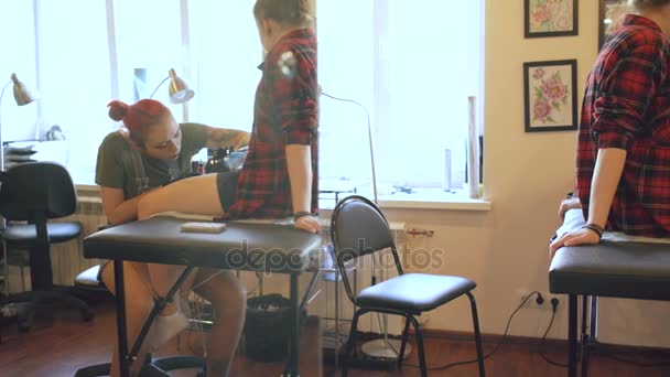 Pięknych czerwonych włosach kobiety artysta tatuażu Tatuowanie zdjęcie na nodze klienta młoda dziewczyna w studio - Materiał filmowy, wideo