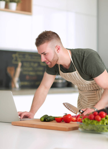 Portrait of man cooking vegetable in the kitchen while looking at a laptop computer on the table - Fotó, kép