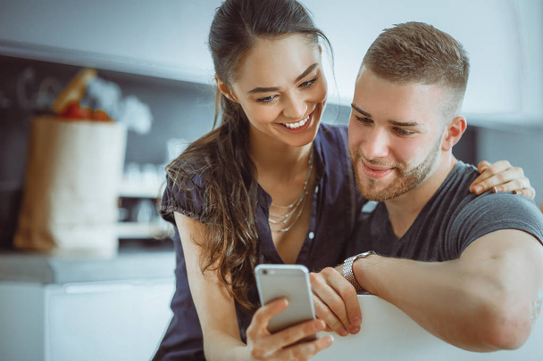 Happy couple using smartphone sitting in kitchen - Foto, afbeelding